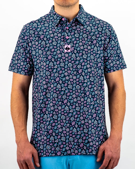 Men’s Daisy Daze golf polo by Pins & Aces featuring an all-over pink, blue, and white daisy floral print on a navy background with an athletic performance fit.
