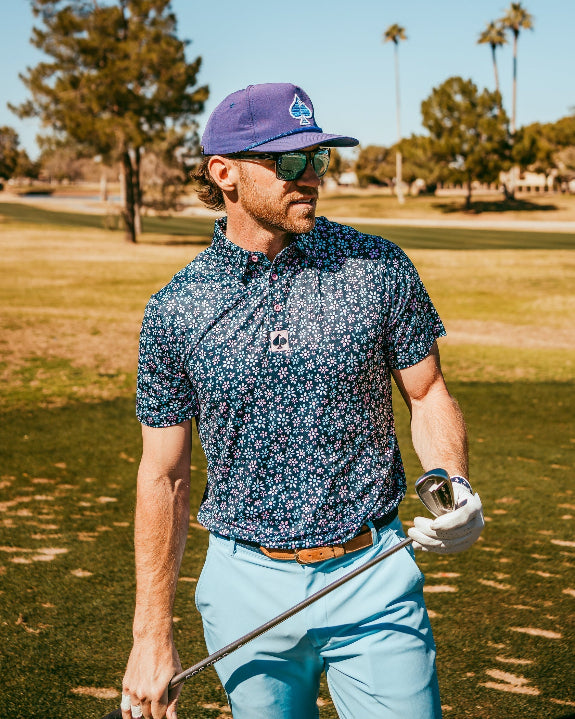 Men’s Daisy Daze golf polo by Pins & Aces featuring an all-over pink, blue, and white daisy floral print on a navy background with an athletic performance fit.