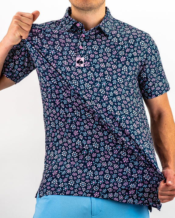 Men’s Daisy Daze golf polo by Pins & Aces featuring an all-over pink, blue, and white daisy floral print on a navy background with an athletic performance fit.