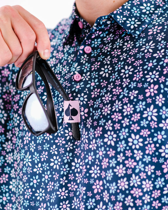Men’s Daisy Daze golf polo by Pins & Aces featuring an all-over pink, blue, and white daisy floral print on a navy background with an athletic performance fit.