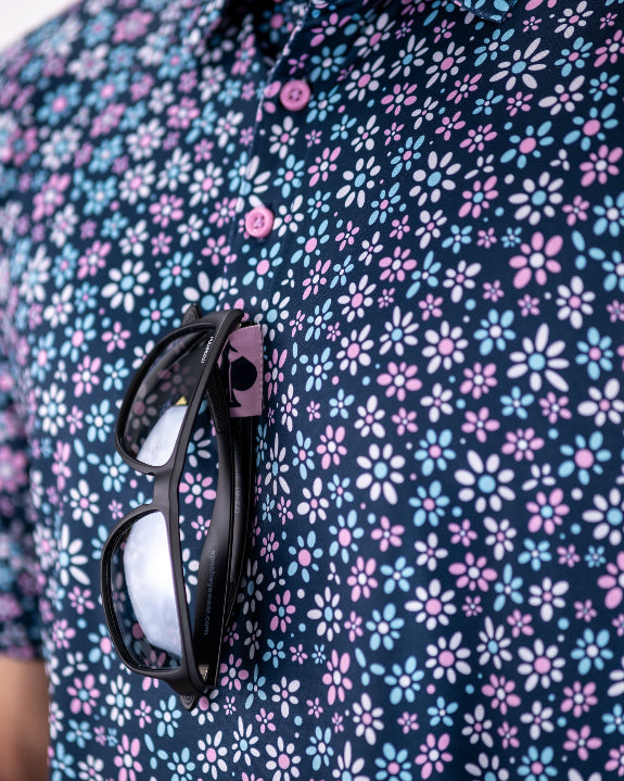 Men’s Daisy Daze golf polo by Pins & Aces featuring an all-over pink, blue, and white daisy floral print on a navy background with an athletic performance fit.