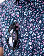 Men’s Daisy Daze golf polo by Pins & Aces featuring an all-over pink, blue, and white daisy floral print on a navy background with an athletic performance fit.