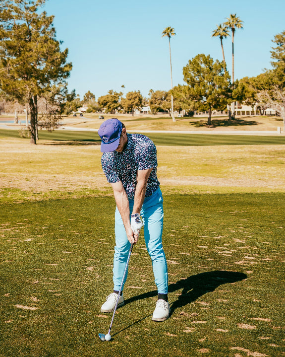 Men’s Daisy Daze golf polo by Pins & Aces featuring an all-over pink, blue, and white daisy floral print on a navy background with an athletic performance fit.