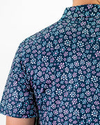 Men’s Daisy Daze golf polo by Pins & Aces featuring an all-over pink, blue, and white daisy floral print on a navy background with an athletic performance fit.