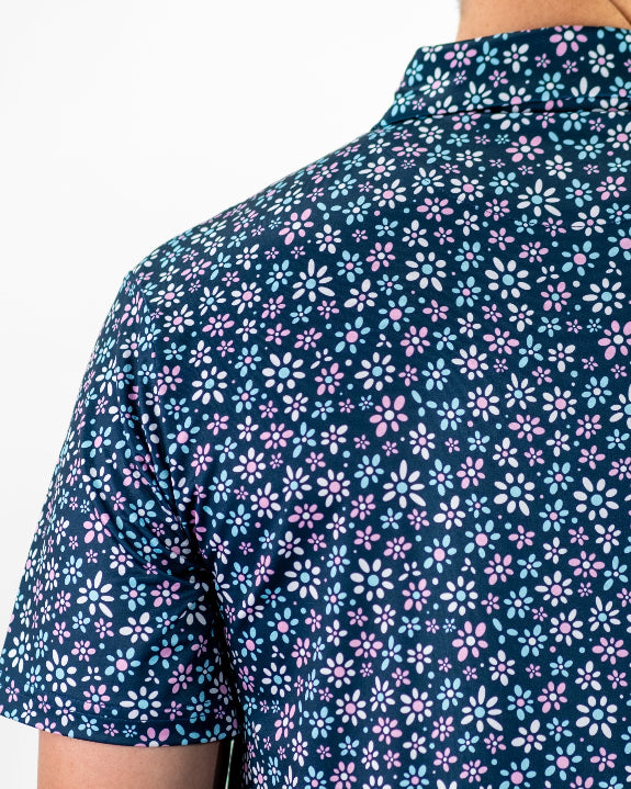 Men’s Daisy Daze golf polo by Pins & Aces featuring an all-over pink, blue, and white daisy floral print on a navy background with an athletic performance fit.