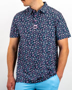 Men’s Daisy Daze golf polo by Pins & Aces featuring an all-over pink, blue, and white daisy floral print on a navy background with an athletic performance fit.