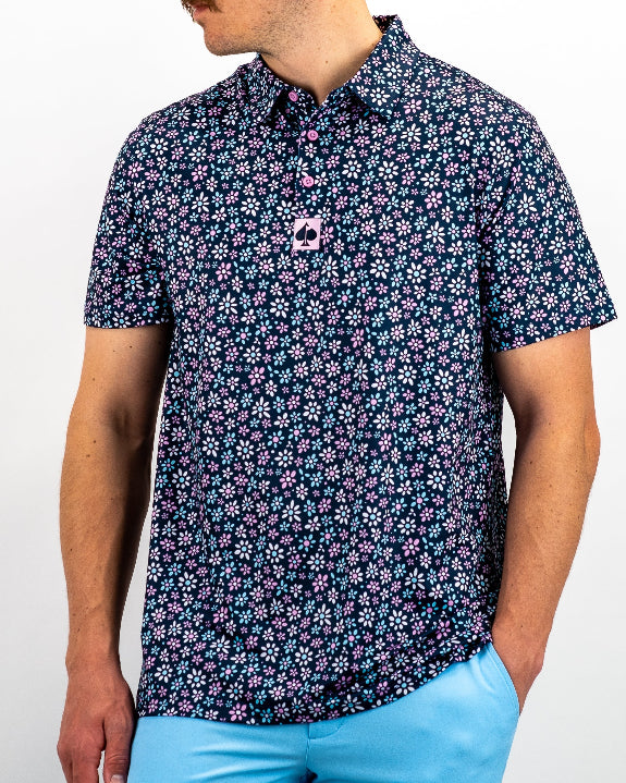Men’s Daisy Daze golf polo by Pins & Aces featuring an all-over pink, blue, and white daisy floral print on a navy background with an athletic performance fit.