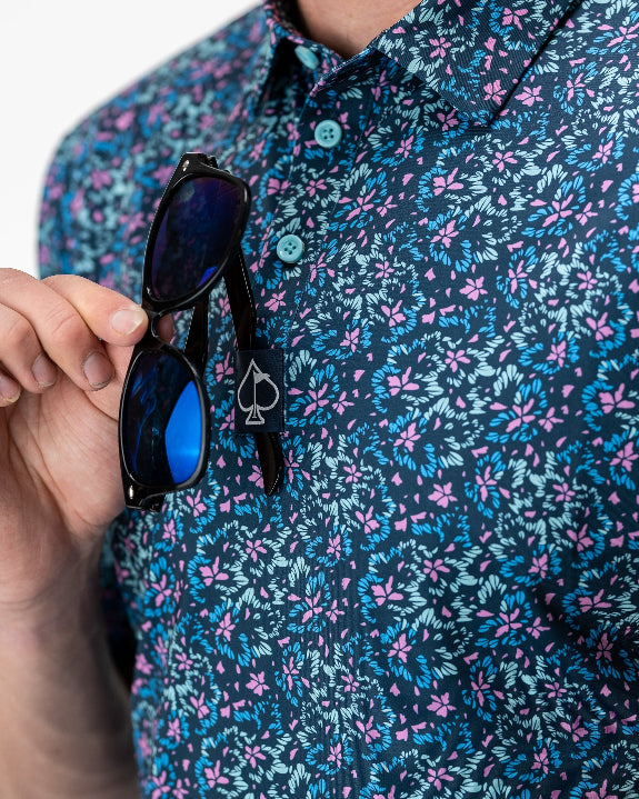 Men’s Fluorescent Floral golf polo by Pins & Aces featuring a vibrant pink, teal, and blue floral pattern on a dark navy short-sleeve performance shirt.