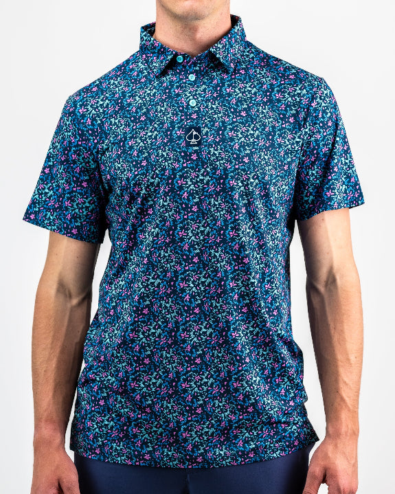 Men’s Fluorescent Floral golf polo by Pins & Aces featuring a vibrant pink, teal, and blue floral pattern on a dark navy short-sleeve performance shirt.