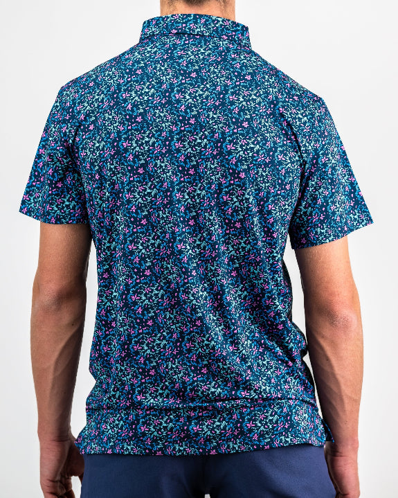 Men’s Fluorescent Floral golf polo by Pins & Aces featuring a vibrant pink, teal, and blue floral pattern on a dark navy short-sleeve performance shirt.