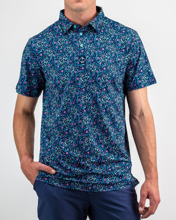Men’s Fluorescent Floral golf polo by Pins & Aces featuring a vibrant pink, teal, and blue floral pattern on a dark navy short-sleeve performance shirt.