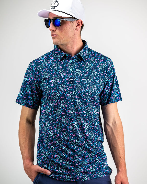Men’s Fluorescent Floral golf polo by Pins & Aces featuring a vibrant pink, teal, and blue floral pattern on a dark navy short-sleeve performance shirt.