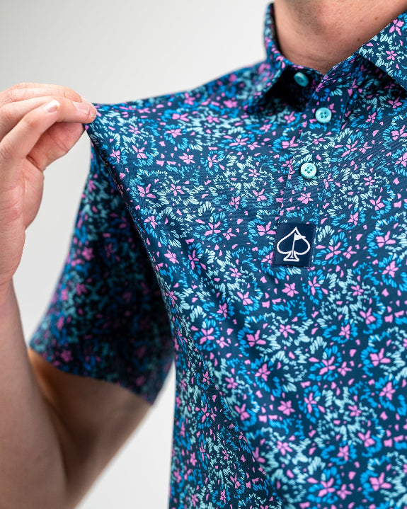 Men’s Fluorescent Floral golf polo by Pins & Aces featuring a vibrant pink, teal, and blue floral pattern on a dark navy short-sleeve performance shirt.
