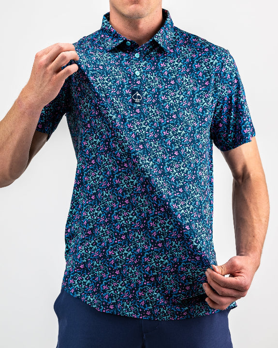 Men’s Fluorescent Floral golf polo by Pins & Aces featuring a vibrant pink, teal, and blue floral pattern on a dark navy short-sleeve performance shirt.