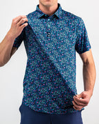 Men’s Fluorescent Floral golf polo by Pins & Aces featuring a vibrant pink, teal, and blue floral pattern on a dark navy short-sleeve performance shirt.