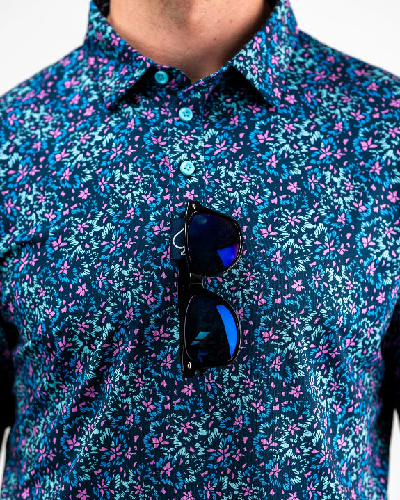 Men’s Fluorescent Floral golf polo by Pins & Aces featuring a vibrant pink, teal, and blue floral pattern on a dark navy short-sleeve performance shirt.