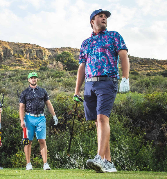 Men’s Cabo Colada golf polo by Pins & Aces featuring a vibrant coral red and aqua tropical floral print on a rich blue background with short sleeves and an athletic performance fit.