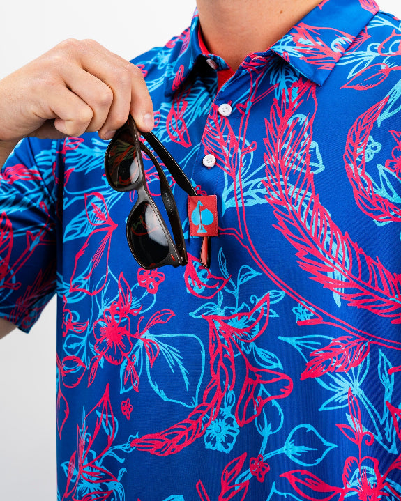 Men’s Cabo Colada golf polo by Pins & Aces featuring a vibrant coral red and aqua tropical floral print on a rich blue background with short sleeves and an athletic performance fit.