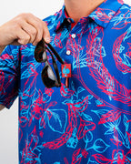 Men’s Cabo Colada golf polo by Pins & Aces featuring a vibrant coral red and aqua tropical floral print on a rich blue background with short sleeves and an athletic performance fit.