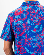 Men’s Cabo Colada golf polo by Pins & Aces featuring a vibrant coral red and aqua tropical floral print on a rich blue background with short sleeves and an athletic performance fit.
