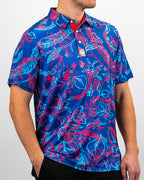 Men’s Cabo Colada golf polo by Pins & Aces featuring a vibrant coral red and aqua tropical floral print on a rich blue background with short sleeves and an athletic performance fit.