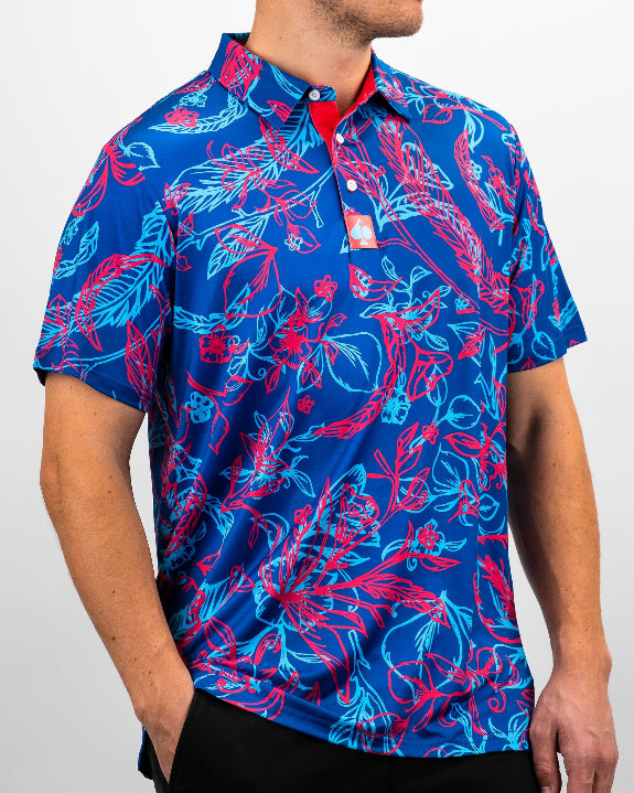 Men’s Cabo Colada golf polo by Pins & Aces featuring a vibrant coral red and aqua tropical floral print on a rich blue background with short sleeves and an athletic performance fit.