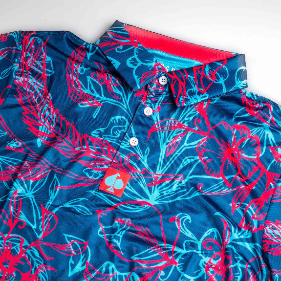 Men’s Cabo Colada golf polo by Pins & Aces featuring a vibrant coral red and aqua tropical floral print on a rich blue background with short sleeves and an athletic performance fit.