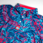 Men’s Cabo Colada golf polo by Pins & Aces featuring a vibrant coral red and aqua tropical floral print on a rich blue background with short sleeves and an athletic performance fit.