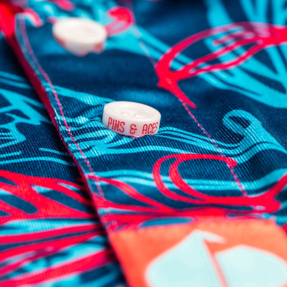 Men’s Cabo Colada golf polo by Pins & Aces featuring a vibrant coral red and aqua tropical floral print on a rich blue background with short sleeves and an athletic performance fit.