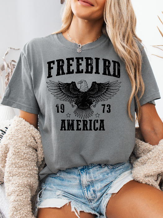 Women’s Freebird 1973 graphic tee featuring a bold Freebird America design, bald eagle graphic, black detailing, and vintage-inspired artwork on a Comfort Colors® cotton T-shirt in Grey