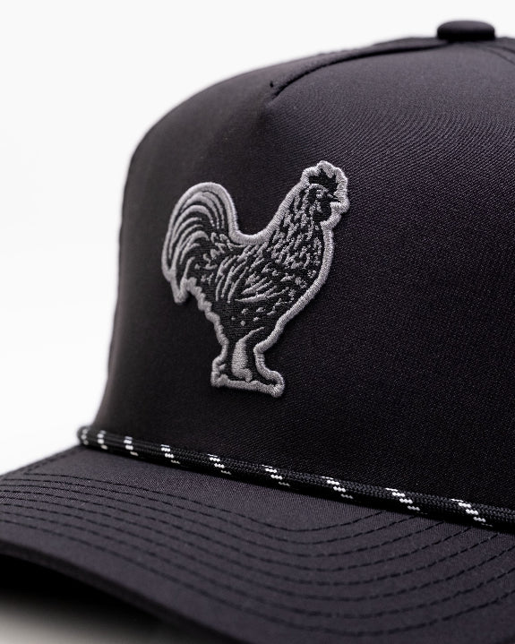 Black Pins & Aces perforated rope golf hat featuring an embroidered rooster on the front, breathable side panels, rope detail, and adjustable snapback design.