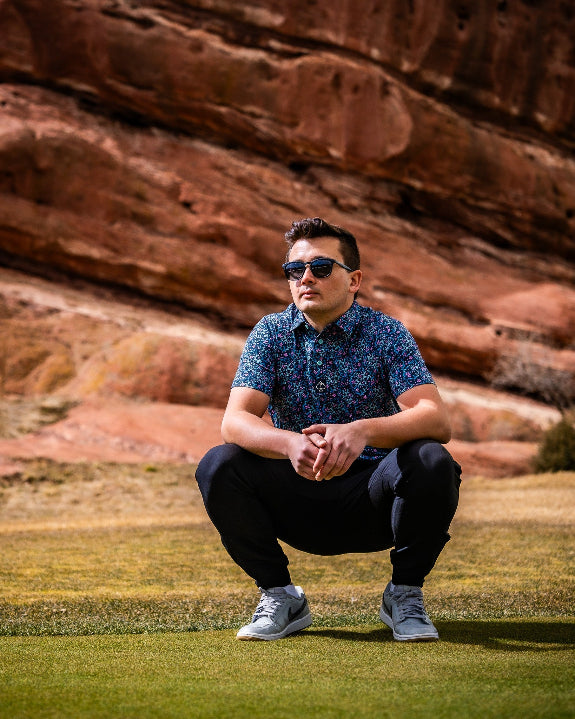Men’s Fluorescent Floral golf polo by Pins & Aces featuring a vibrant pink, teal, and blue floral pattern on a dark navy short-sleeve performance shirt.