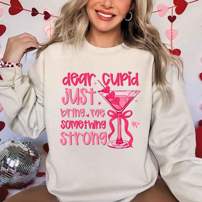 Dear Cupid Something Strong Valentine’s Day crewneck sweatshirt featuring a funny Cupid-inspired graphic on a cozy relaxed-fit unisex sweatshirt in Sand