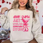 Dear Cupid Something Strong Valentine’s Day crewneck sweatshirt featuring a funny Cupid-inspired graphic on a cozy relaxed-fit unisex sweatshirt in Sand