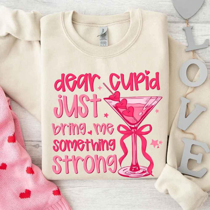 Dear Cupid Something Strong Valentine’s Day crewneck sweatshirt featuring a funny Cupid-inspired graphic on a cozy relaxed-fit unisex sweatshirt in Sand