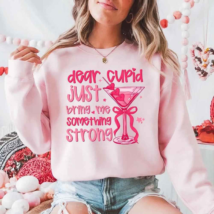 Dear Cupid Something Strong Valentine’s Day crewneck sweatshirt featuring a funny Cupid-inspired graphic on a cozy relaxed-fit unisex sweatshirt in Light Pink