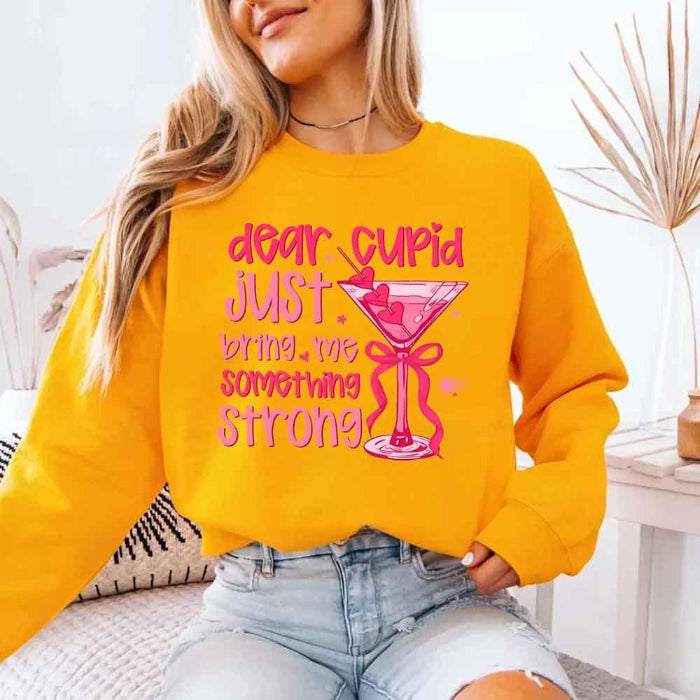 Dear Cupid Something Strong Valentine’s Day crewneck sweatshirt featuring a funny Cupid-inspired graphic on a cozy relaxed-fit unisex sweatshirt in Gold