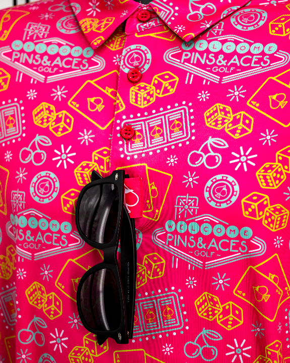 Men’s Big Ace Fuchsia golf polo by Pins & Aces featuring a bright pink casino print with playing cards, dice, poker chips, slot machines, and an athletic performance fit.
