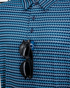 Men’s Rising Tides golf polo by Pins & Aces featuring a repeating blue and navy wave-inspired pattern, short sleeves, and an athletic performance fit.