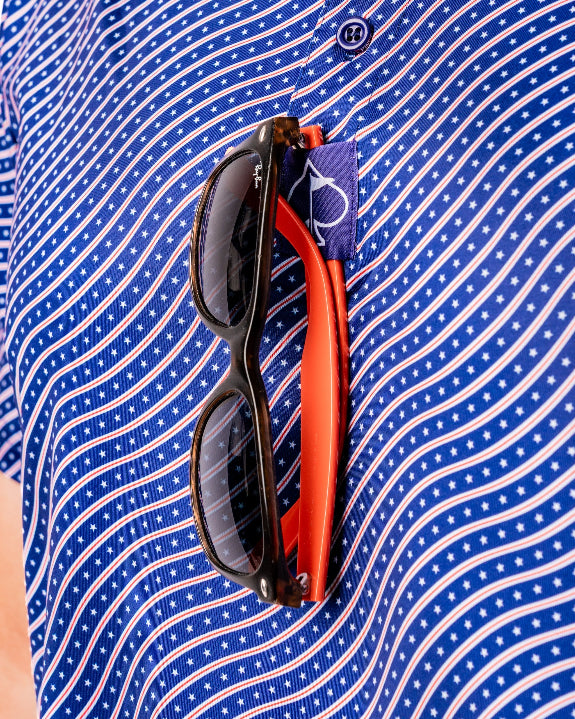 Men’s Wavy Stars & Stripes golf polo by Pins & Aces featuring a navy, red, and white wavy patriotic pattern with short sleeves and an athletic performance fit.