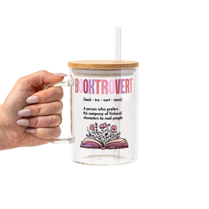 Clear glass cup with bamboo lid and straw featuring pink Booktrovert lettering, a definition about preferring fictional characters to real people, and an open book filled with pink and purple flowers.