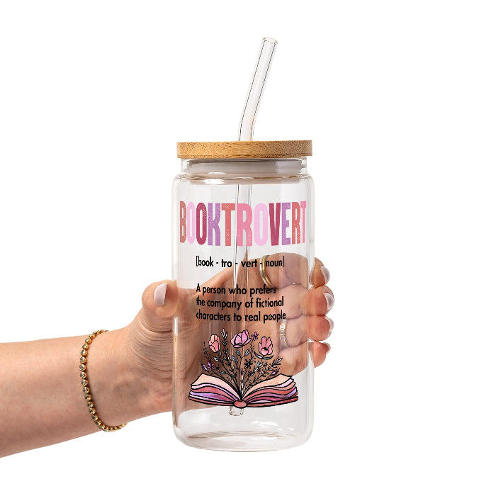 Clear glass cup with bamboo lid and straw featuring pink Booktrovert lettering, a definition about preferring fictional characters to real people, and an open book filled with pink and purple flowers.