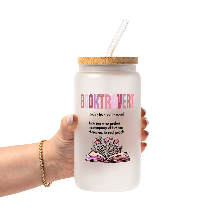 Clear glass cup with bamboo lid and straw featuring pink Booktrovert lettering, a definition about preferring fictional characters to real people, and an open book filled with pink and purple flowers.