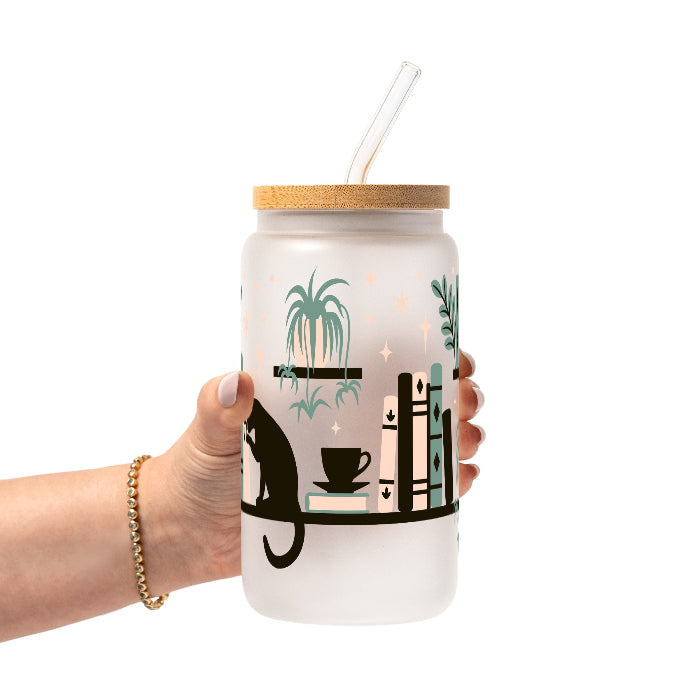 Bookish drinkware featuring a cozy wraparound bookshelf design with books, a curious kitty, and trailing green plants, available in glass cups, tumblers, and ceramic mugs.