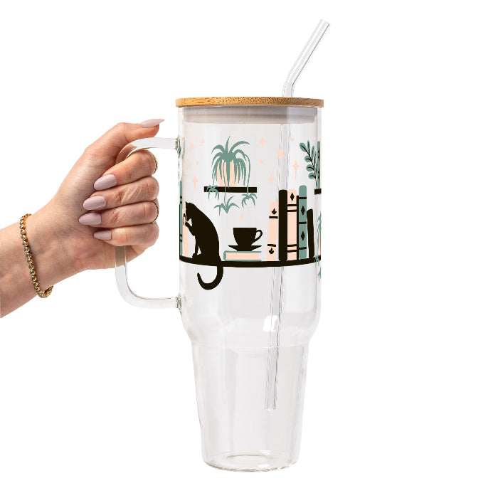 Bookish drinkware featuring a cozy wraparound bookshelf design with books, a curious kitty, and trailing green plants, available in glass cups, tumblers, and ceramic mugs.