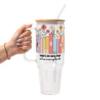 Clear glass cup with bamboo lid and straw featuring colorful pastel books surrounded by pink, yellow and white wildflowers with “There’s No Such Thing as Too Many Books” lettering.