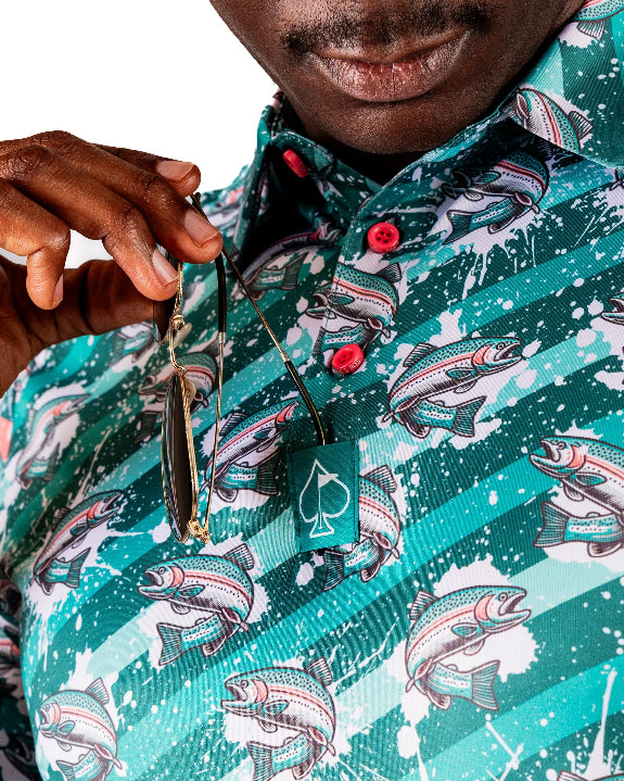 Men’s Trout Splatter golf polo by Pins & Aces featuring an all-over trout fishing print with teal, aqua, coral, and white splatter details and an athletic performance fit.
