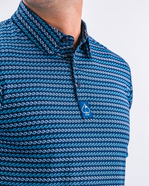 Men’s Rising Tides golf polo by Pins & Aces featuring a repeating blue and navy wave-inspired pattern, short sleeves, and an athletic performance fit.