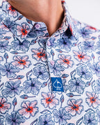 Men’s Hibiscus Harmony golf polo by Pins & Aces featuring blue and coral hibiscus flowers on a white lightweight performance golf shirt.