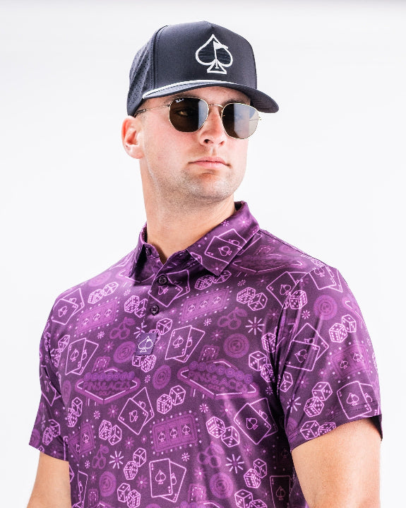 Men’s purple casino golf polo by Pins & Aces featuring an all-over pattern of playing cards, dice, poker chips, slot machines, and Las Vegas-inspired graphics.